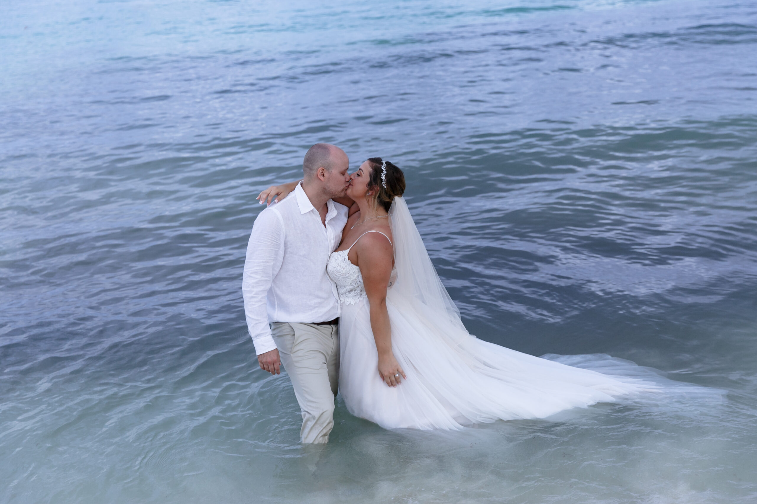 Newlyweds kiss, standing in the ocean with her dress floating in the water