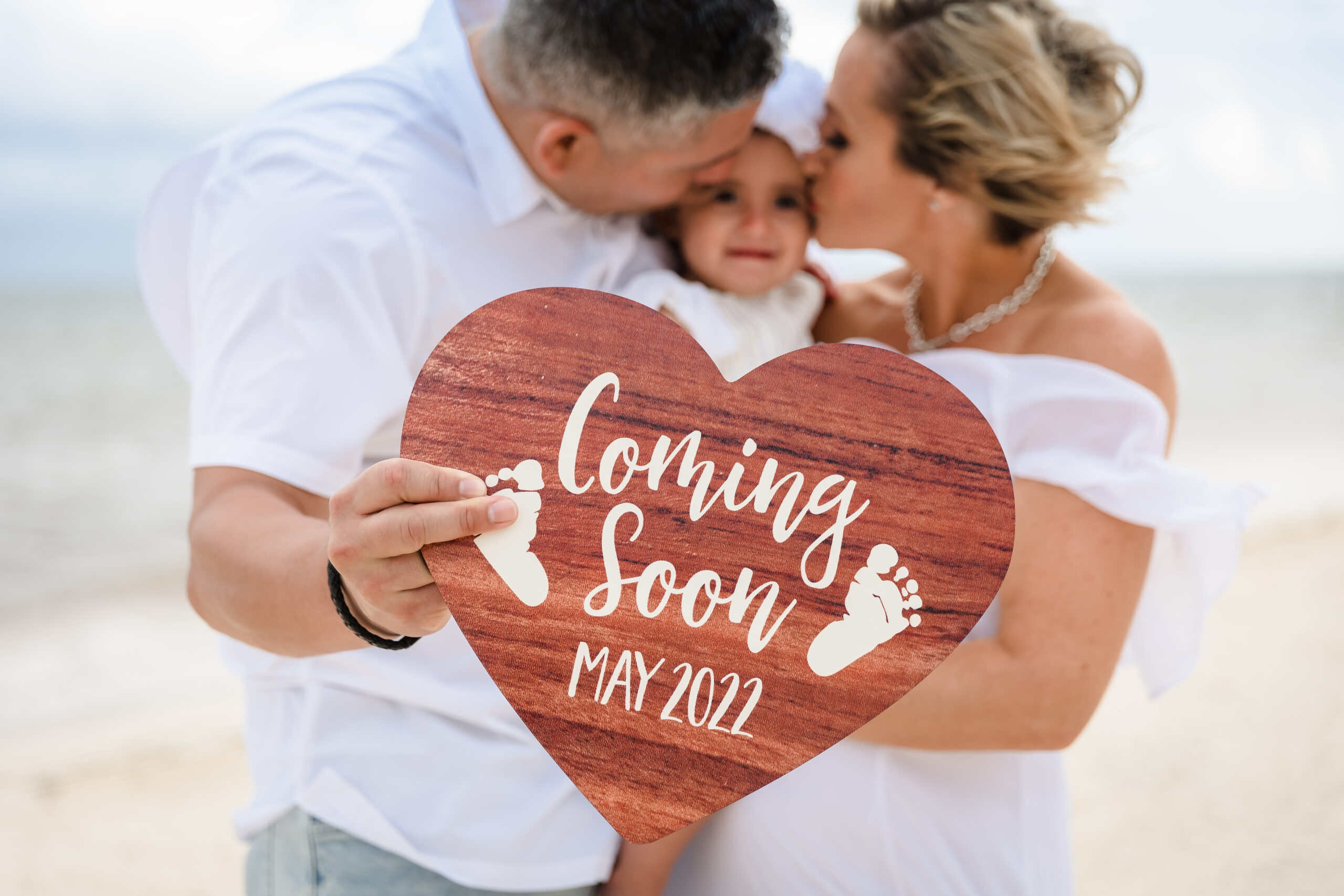 Expectant couple kiss their first child while holding a "coming soon" sign