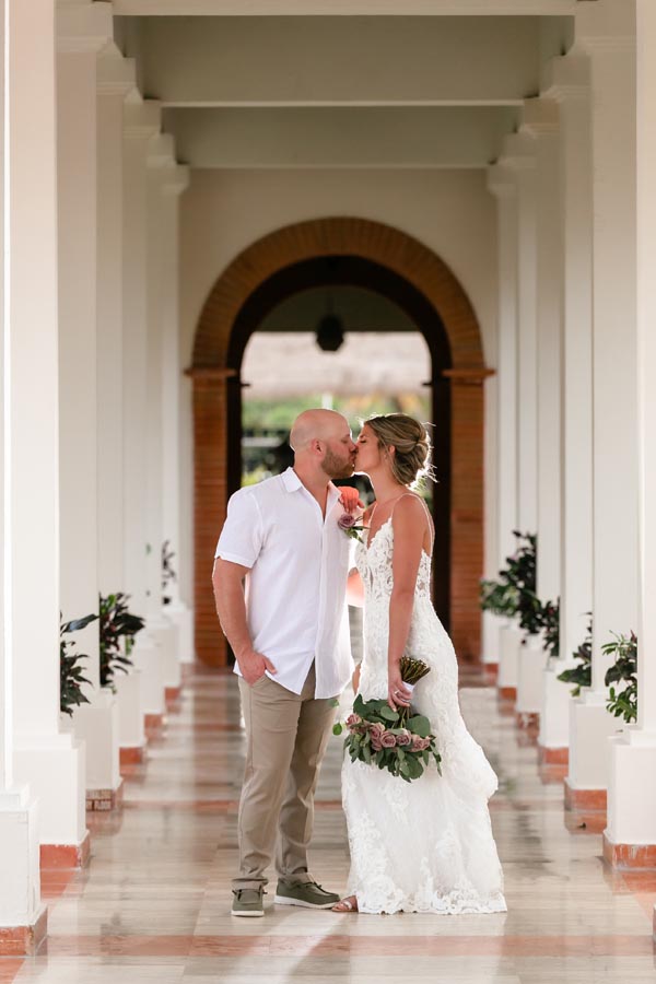 Newlyweds kiss in an arched hallway lined with columns