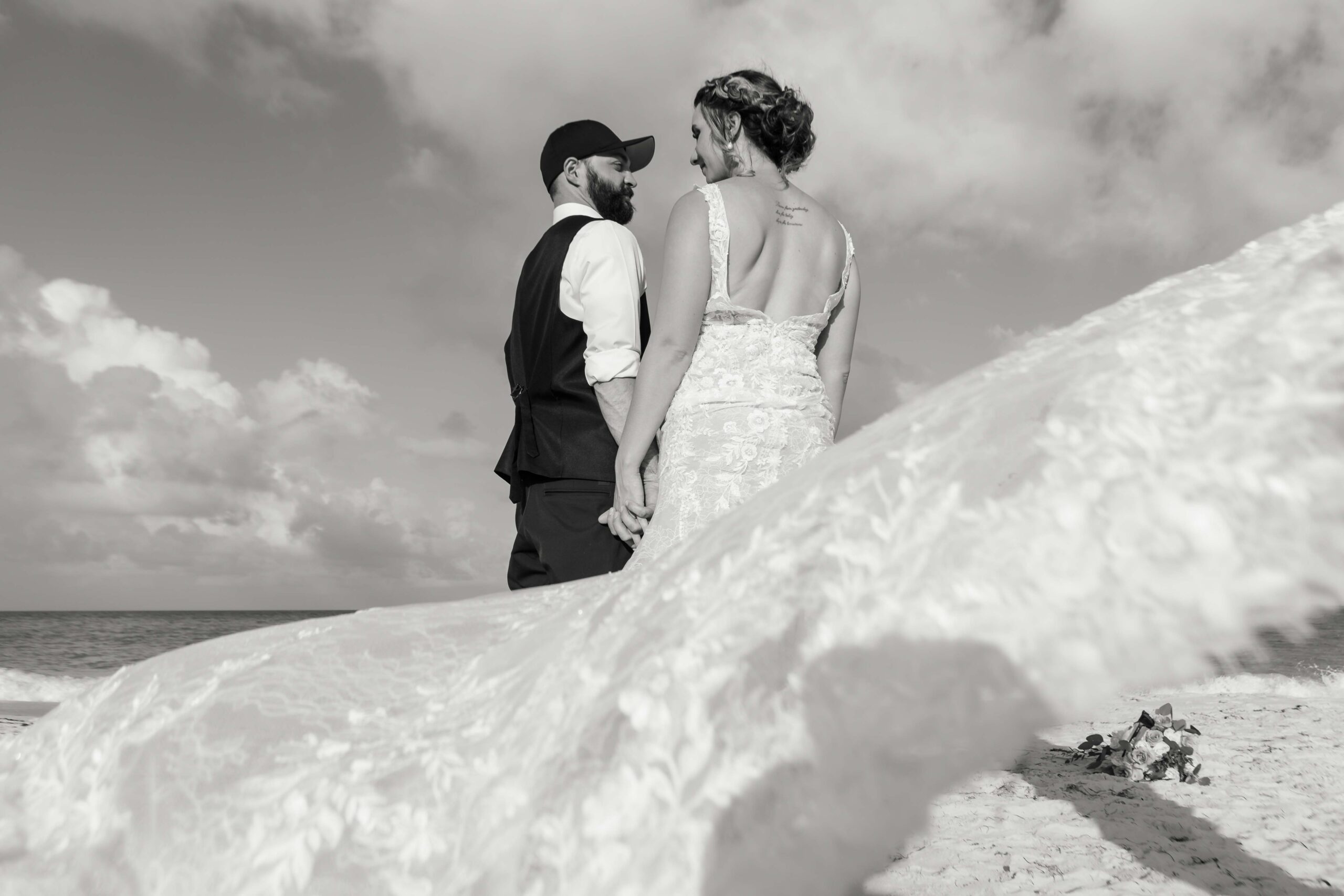 Wind billows brides lace train as newlyweds stroll along the beach