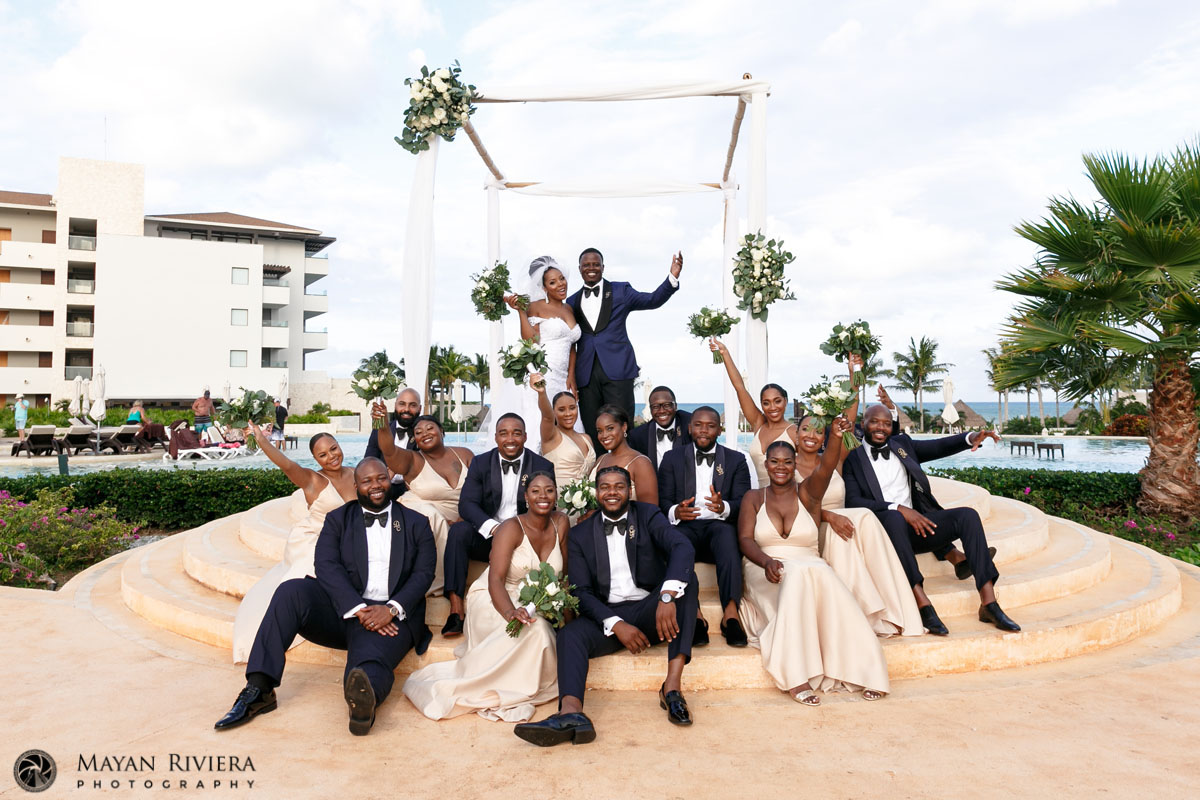 Large Wedding Party Sits On Steps In Front of Cheering Newlyweds
