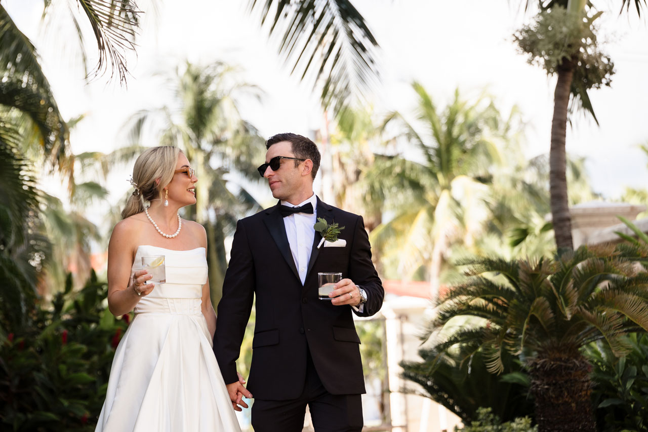 Relaxed newlyweds stroll through gardens with beverage