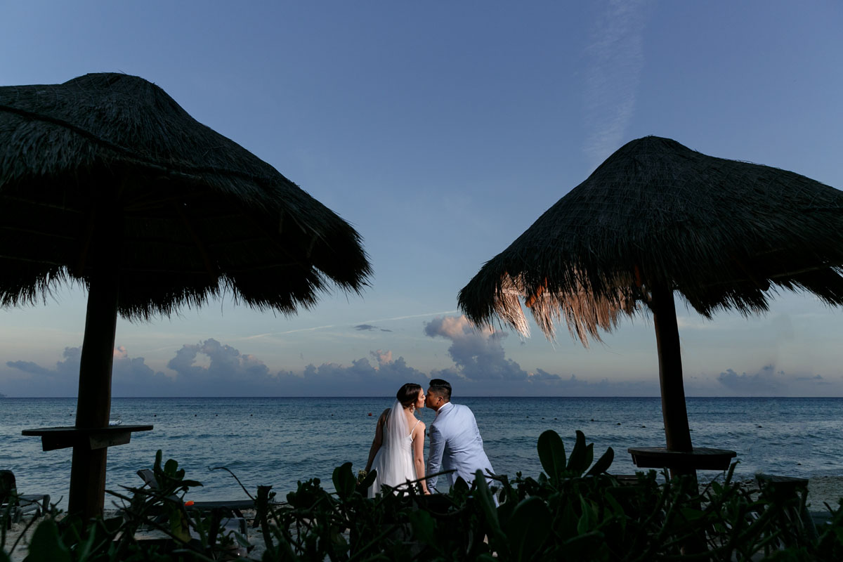 NewlywedsKissByBeachsidePalapas