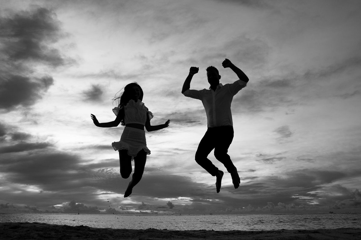 NewlyEngagedCoupleJumpForJoyOnPdCBeachAtSunrise