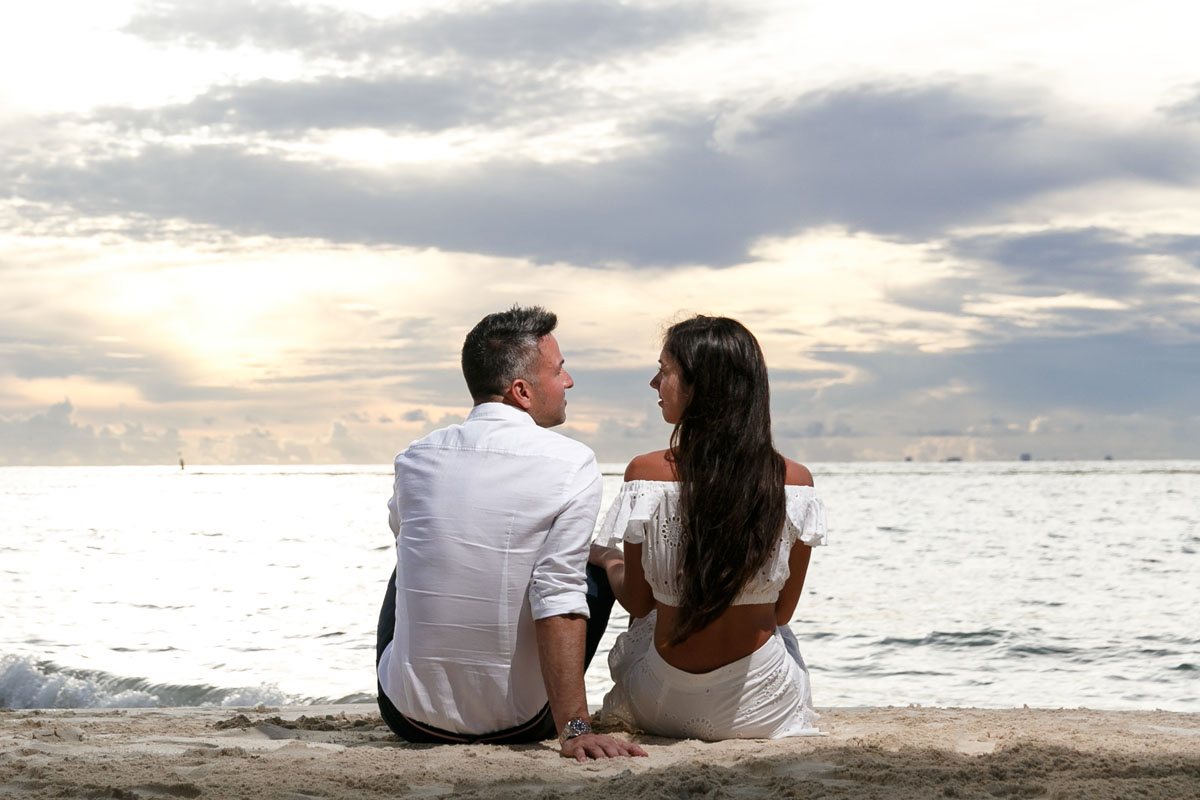 NewlyEngagedCoupleSitOnPdCBeachAtSunrise