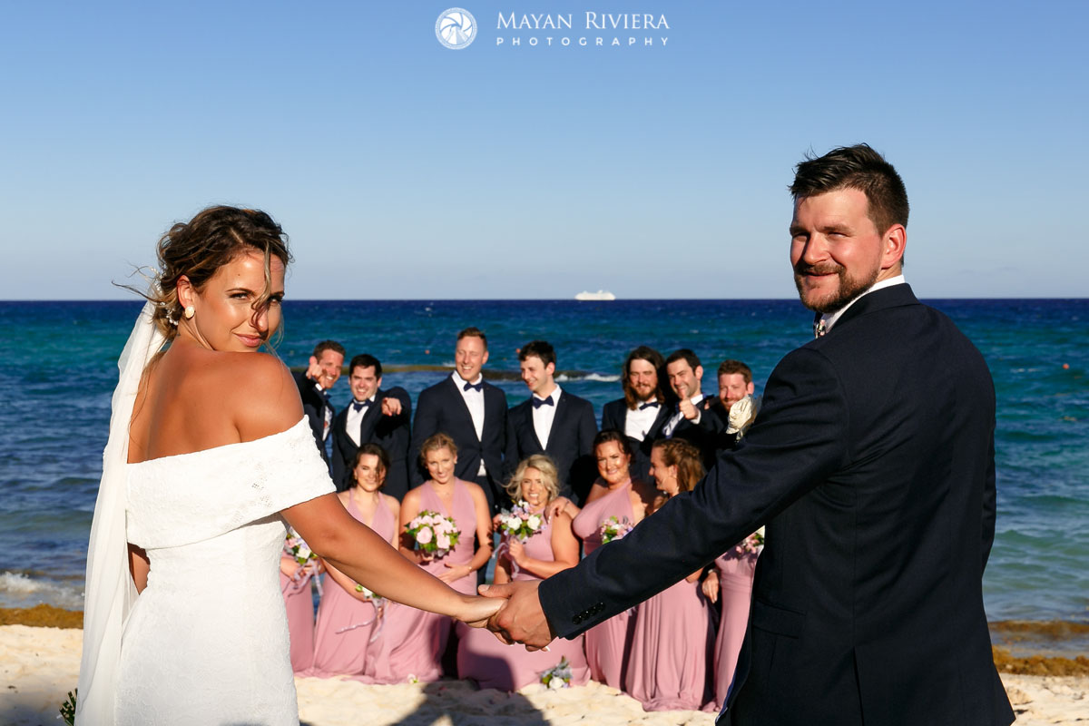 Newlyweds smile over their shoulders with wedding party in the background on the beach