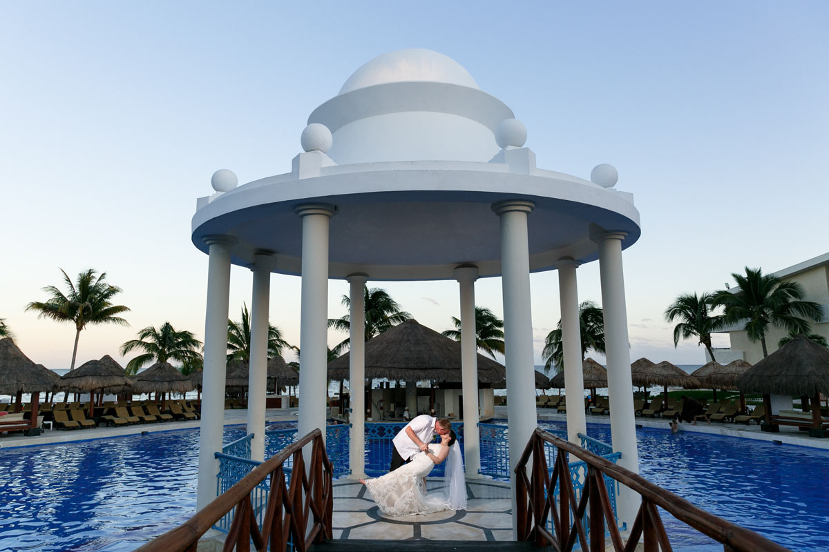 NewlywedsEmbraceInDipUnderDomePavillion