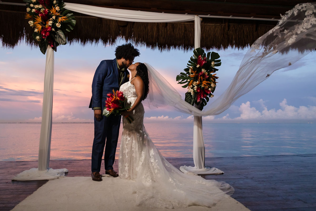 Newlyweds kiss on pier silhouetted by magical sunset
