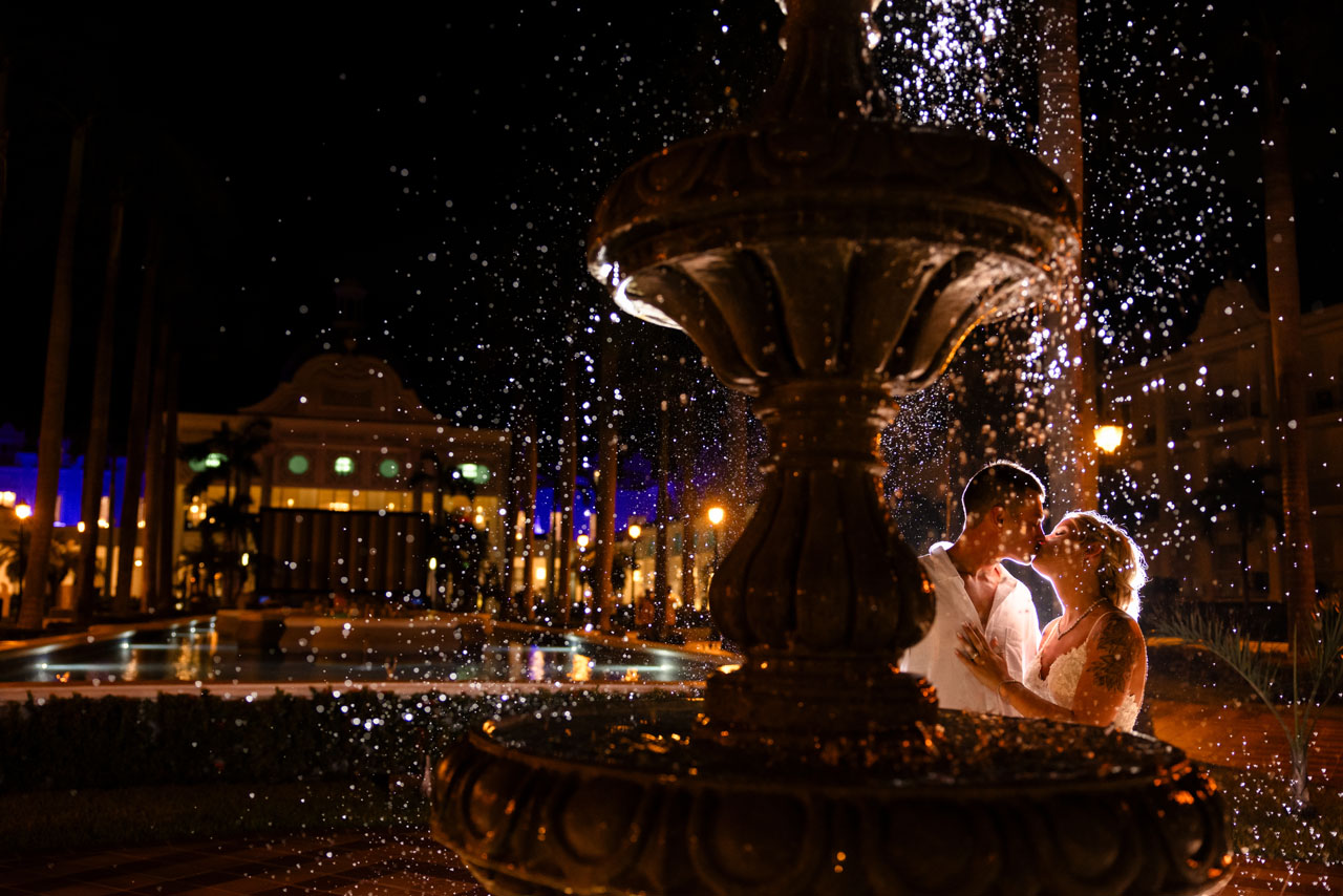 Newlyweds kiss behind spray of water fountain lighting up the night