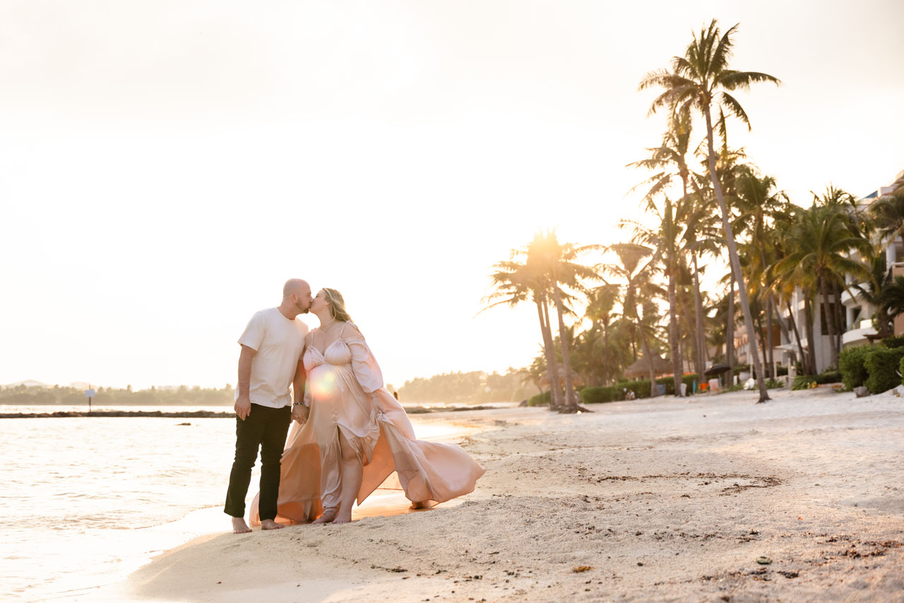 Expectant parents kiss on beach with a sunbeam shining on her baby bump