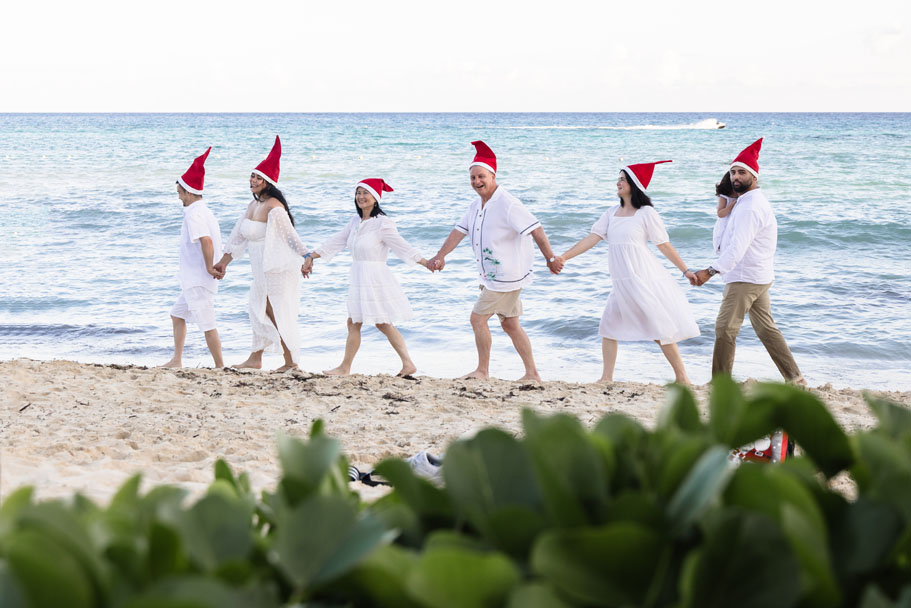 Happy family strolls the beach in a line holding hands, wearing Santa hats
